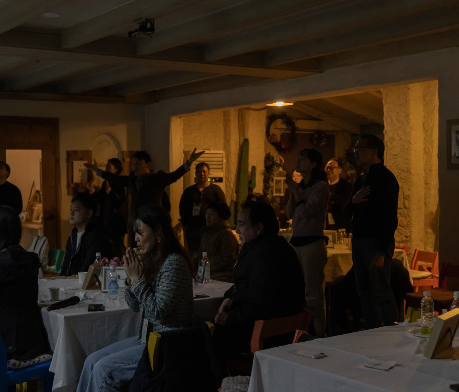 Leaders worshipping together in a warm, lamplit room — one praying with hands clasped, others with hands raised